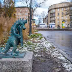 Mały smok z brązu, symbol smoczy szlak kraków, stoi na kamiennym postumencie. W tle miejski krajobraz z budynkami i mokrą nawierzchnią.