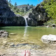 Pies descalzos en la orilla del **barranco de la Encantada**, con una cascada cayendo en una poza de agua verde rodeada de rocas y vegetación.