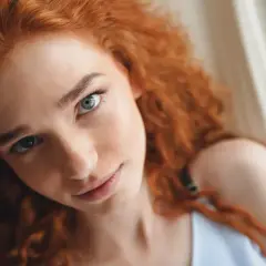 Primer plano de una joven con cabello pelirrojo natural y ojos azules, recostada en una hamaca.