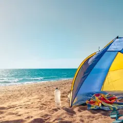 Kolorowy namiot na plaży, obok butelka wody, ręcznik i klapki. Słońce odbija się w morzu.