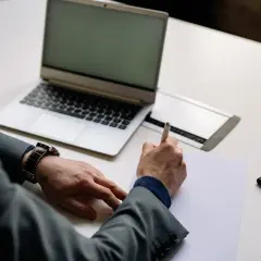 Mężczyzna w garniturze pisze na kartce, obok laptop, telefon i zegarek. Czas na rozliczenie vat-z.