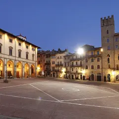 Piazza illuminata ad Arezzo, con portici, edifici storici e una torre. Sembra proprio che la vita è bella ad Arezzo.