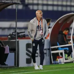 Łysy mężczyzna w okularach, trener reprezentacji Hiszpanii, w szarej kurtce i granatowych spodniach, stoi na boisku.
