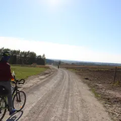 Rowerzysta na szutrowej drodze, która wydaje się być częścią najdłuższej trasy rowerowej w Polsce. Słońce świeci na błękitnym niebie.