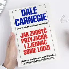 Książka "Jak zdobyć przyjaciół i zjednać sobie ludzi" autorstwa Dale'a Carnegie, trzymana w dłoni, leży obok klawiatury i rośliny.