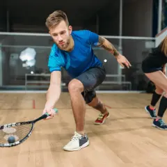 Mężczyzna w niebieskiej koszulce i szarych spodenkach, z rakietą do squasha, pokazuje, jakie buty do squasha są najlepsze do dynamicznej gry. Obok kobieta w czarnej sukience.