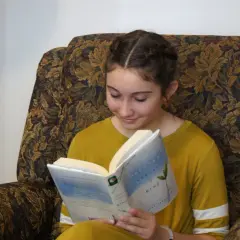Dziewczyna w warkoczach czyta książkę "Between Shades of Gray", idealną dla 14 latki. Siedzi w wygodnym, wzorzystym fotelu.