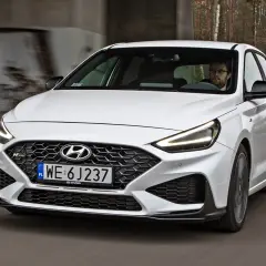 Biały Hyundai i30 N z dynamicznym grillem i agresywnym zderzakiem jedzie po drodze wśród drzew.