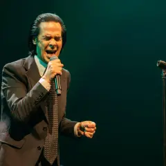 Nick Cave z pasją wykonuje swoje utwory, śpiewając do mikrofonu w eleganckim garniturze.