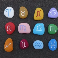 Kolorowe kamienie z symbolami znaków zodiaku po kolei, ułożone na ciemnym piasku.