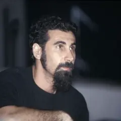 Serj Tankian, wokalista System of a Down, z charakterystyczną brodą i słuchawką w uchu, gotowy do wykonania kolejnych energetycznych utworów.
