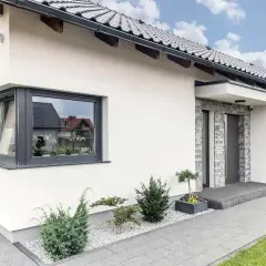 Nowoczesny dom z białym tynkiem, szarym dachem i efektownym **oknem narożnym ze słupkiem**. Przed wejściem ozdobne krzewy i kamyczki.