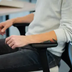 Osoba z tatuażem na ramieniu siedzi w biurowym fotelu, co może symbolizować siedzący tryb życia.