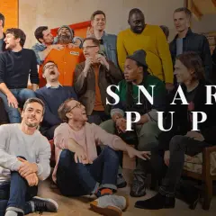 Grupa ludzi, w tym mężczyzna w pomarańczowym kombinezonie, pozuje do zdjęcia z napisem "SNARKY PUPPY".
