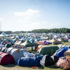 Ein Meer von Zelten beim Roskilde Festival. Menschen entspannen sich im Freien unter blauem Himmel, umgeben von bunten Unterkünften.