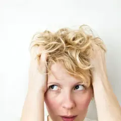 Kobieta z blond włosami, trzymająca się za głowę, z wyrazem twarzy sugerującym wahania nastroju i płaczliwość.