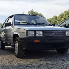 Srebrny Renault 11 z lat 80. stoi na żwirowym podjeździe, obok niebieskiego beczki i innego samochodu.