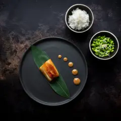 A perfectly cooked nobu miso black cod rests on a dark plate with a green leaf. Drizzled sauce and sides of rice and edamame complete the elegant dish.