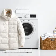 Biała kurtka puchowa wisi obok pralki, gotowa do prania. Obok koszyk z ręcznikami i płynem do prania, idealny do tego, jak prać kurtkę puchową.