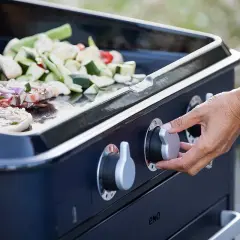Main d'une personne tournant le bouton pour allumer plancha gaz. Viande et légumes coupés sont prêts à cuire sur la plaque.