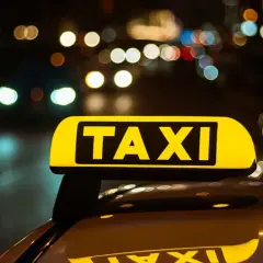 Żółty znak TAXI na dachu samochodu nocą. W tle rozmyte światła miasta, sugerujące licencję taxi w ruchu.