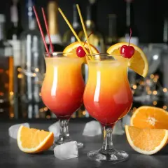 Dwa drinki Tequila Sunrise, ozdobione pomarańczą i wiśnią, stoją na barze. W tle widać butelki alkoholu i migoczące światełka. Idealne na wieczór z przyjaciółmi, jak w najlepszych lokalach Baczewski.
