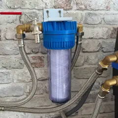 Filtre à eau avec cartouche visible, raccordé à un adoucisseur qui coule en permanence. Tuyaux flexibles et robinet rouge.