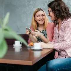 Dwie kobiety rozmawiają przy kawie, tworząc intymne tête-à-tête. Jedna pokazuje coś na telefonie, druga uśmiecha się.