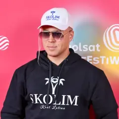 Skolim, Król Latino, w czarnej bluzie z kapturem i białej czapce, emanuje barwami szczęścia na tle festiwalowej ścianki.