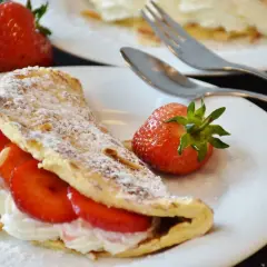 Puszysty omlet z bitą śmietaną i truskawkami, posypany cukrem pudrem i płatkami migdałów. Obok leży świeża truskawka.