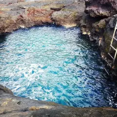 Un charco de los abrigos natural, con aguas cristalinas y una escalera metálica para acceder. Rodeado de rocas volcánicas.