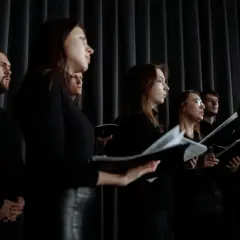 Chór śpiewa, prezentując różne rodzaje głosów męskich. Widać mężczyzn i kobiety w czarnych strojach, trzymających nuty.