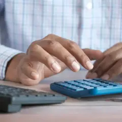 Hände tippen auf einem blauen Taschenrechner, während eine gelbe Tasse und eine Tastatur daneben stehen. Ein Mann berechnet sein business administration gehalt.