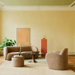 Ein Wohnzimmer mit gelben Wänden und grüner Decke. Ein beiger Teddy-Sofa und Sessel laden zum Entspannen ein. Ein Kunstwerk in Orange und ein Couchtisch mit Glasplatte runden das Bild ab. So kann man richtig streichen mit Rolle.