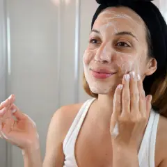 Mujer con diadema negra aplica espuma en su rostro, parte de su rutina de limpieza facial.