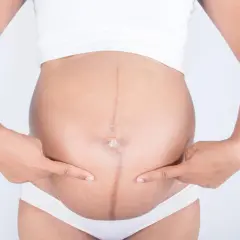Ciężarna kobieta wskazuje palcami na ciemną linię biegnącą przez środek brzucha, zwaną linea negra.