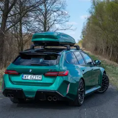 Zielone BMW M5 Touring z boxem dachowym na tle drzew i nieba.