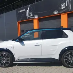Biały Alpine A290 z niebieskimi akcentami stoi przed budynkiem z pomarańczowymi drzwiami garażowymi.