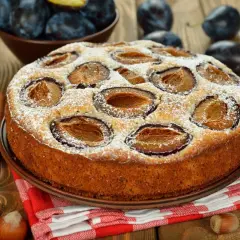 Pyszne ciasto ze śliwkami, posypane cukrem pudrem, na drewnianym blacie. Obok świeże śliwki, orzechy i laski cynamonu.
