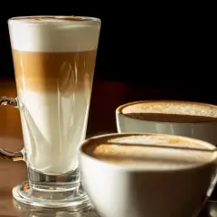 Kawa biała w wysokiej szklance z warstwami mleka i espresso, obok trzy filiżanki kawy z pianką.