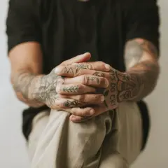 Dłonie z tatuażem handpoke, ozdobione wzorami i napisem "Boys Do Cry", splecione na kolanach.