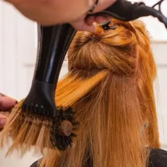 Manos con tatuajes secan y peinan cabello pelirrojo con secador y cepillo redondo. El brushing que es, es una técnica de estilismo.