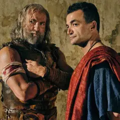 Två män i antika kläder, en med skägg och läderrustning, den andra i röd mantel, står nära varandra. Kanske en scen från Spartacus: House of Ashur.