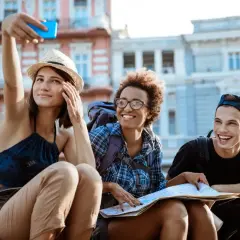 Tres amigos sonríen mientras se toman una selfie en la calle, planeando cómo conocer gente para viajar y explorar la ciudad.