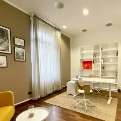 Arredamento studio psicologo: scrivania bianca, libreria, sedie, poltrona gialla, quadri e tenda.
