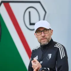 Nowy trener Legii w białej czapce i okularach, na tle klubowego herbu.