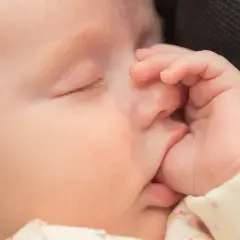 Urocze ssanie kciuka u niemowlaka, który zasypia. Dłoń delikatnie dotyka twarzy, a oczy są zamknięte.