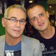 Dwóch mężczyzn pozuje do zdjęcia. Starszy, w okularach i jasnej marynarce, zastanawia się, czy prowadzący naprawdę zna wszystkie piosenki, czy korzysta z promptera.