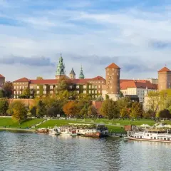 Malownicze miasto nad Wisłą z Wawelem w jesiennej odsłonie. Statki wycieczkowe cumują przy brzegu, a na trawniku odpoczywają spacerowicze.