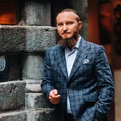 Elegancki mężczyzna w garniturze w kratę, z brodą, oparty o kamienną ścianę. Idealny przykład, jaki krawat do garnituru w kratę wybrać.
